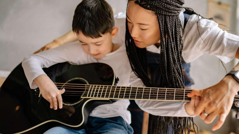 young boy playing black guitar with assistance from teacher