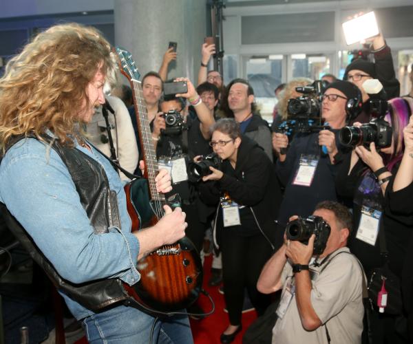 Performer playing electric guitar in front of group of media photographers with cameras out and visible camera flashes